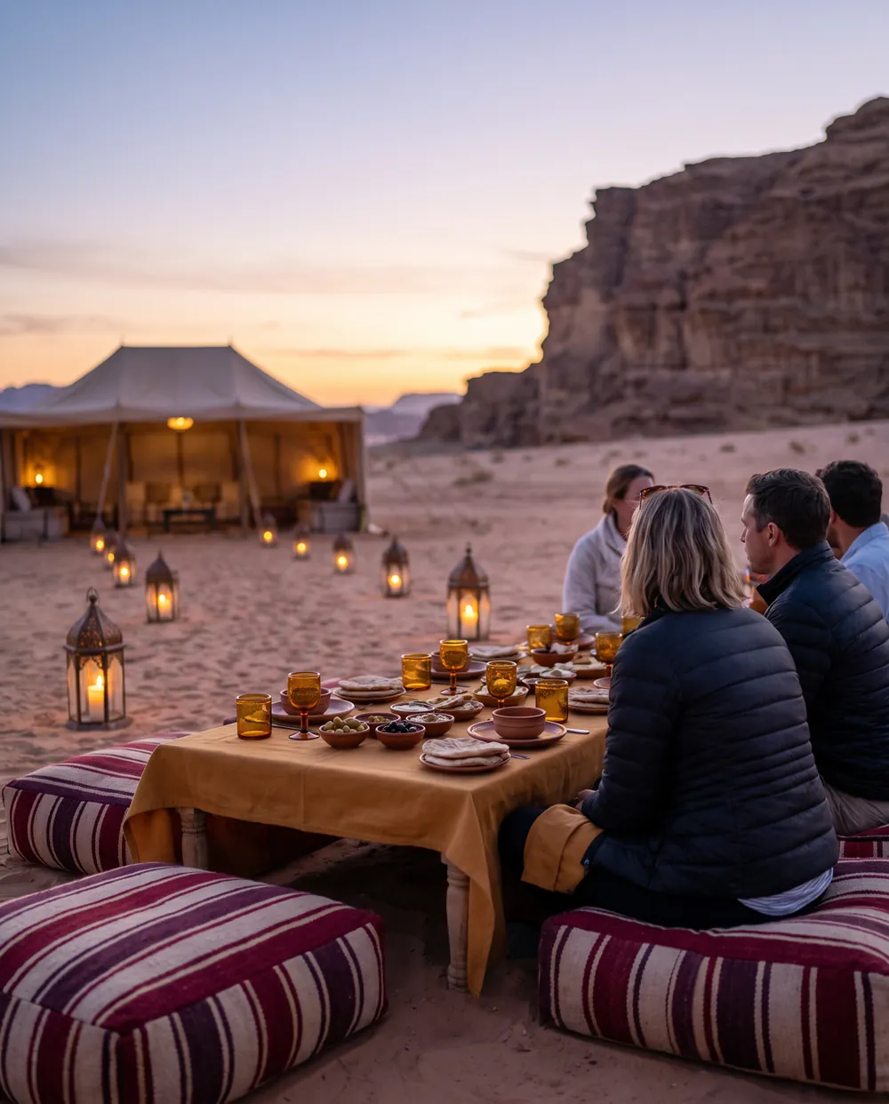 Un dîner dressé à même le sable au crépuscule, entouré de lanternes ajourées, avec une tente éclairée au loin et une falaise de grès.