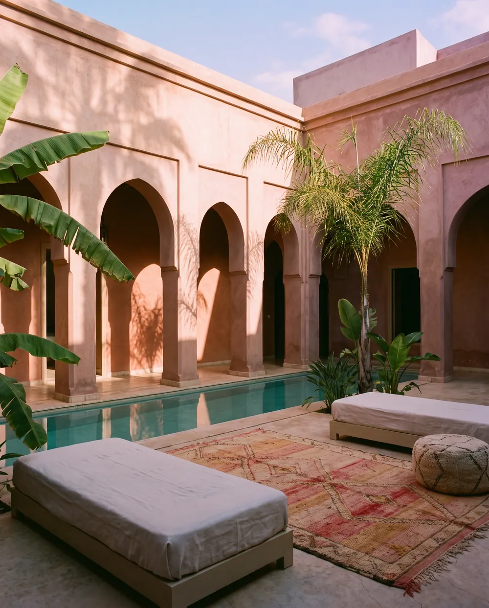 La cour d’un riad au tadelakt rose, ses arches en fer à cheval et un long bassin turquoise, avec deux lits bas et un tapis Beni Ourain.